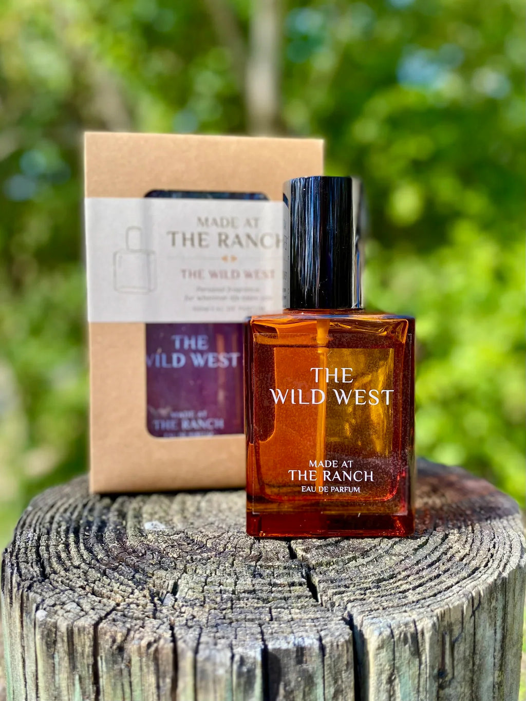 Perfume bottle labeled 'The Wild West' by 'Made at The Ranch' on a wooden stump with a blurred green background.