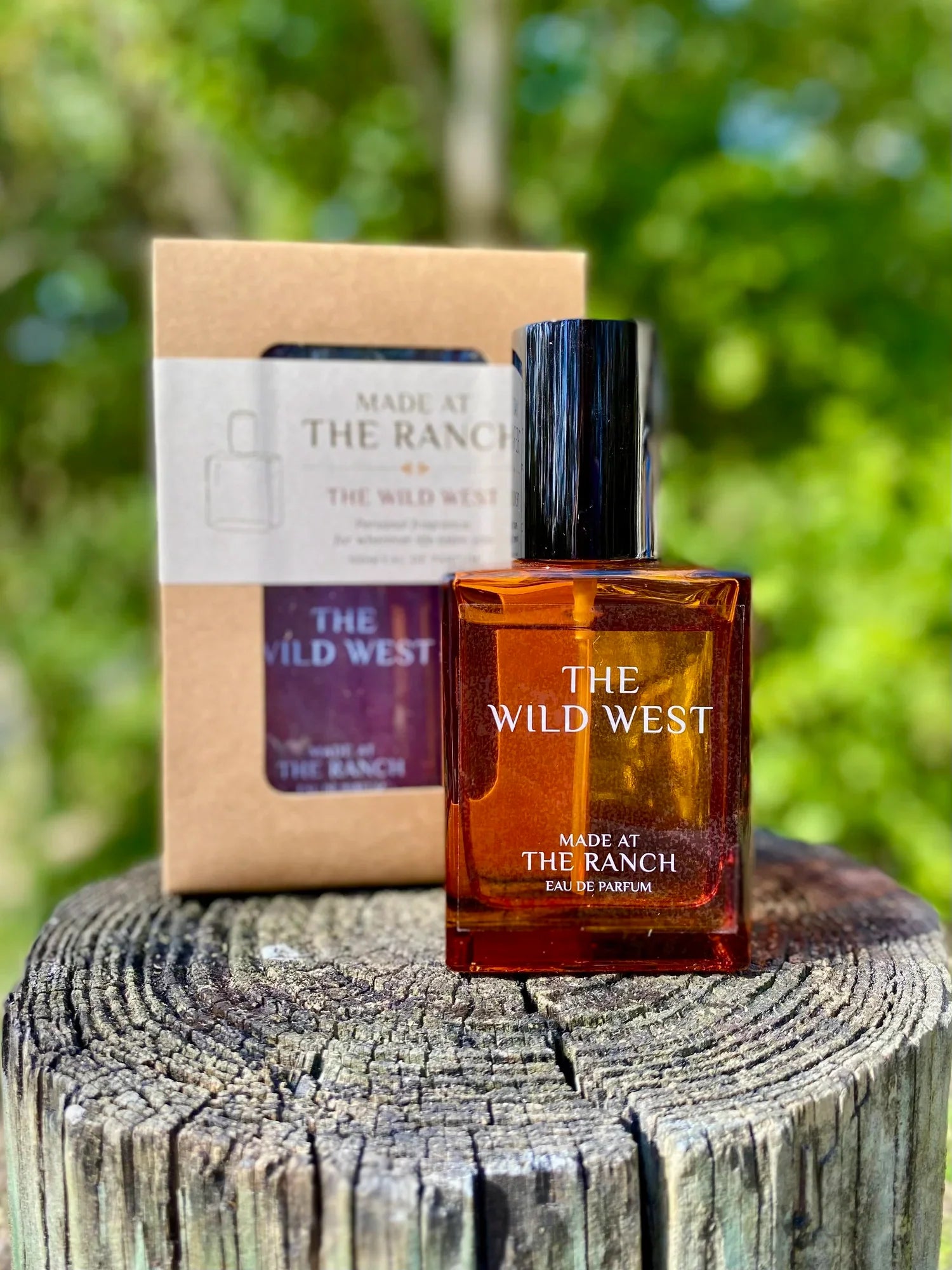 Perfume bottle labeled 'The Wild West' by 'Made at The Ranch' on a wooden stump with a blurred green background.